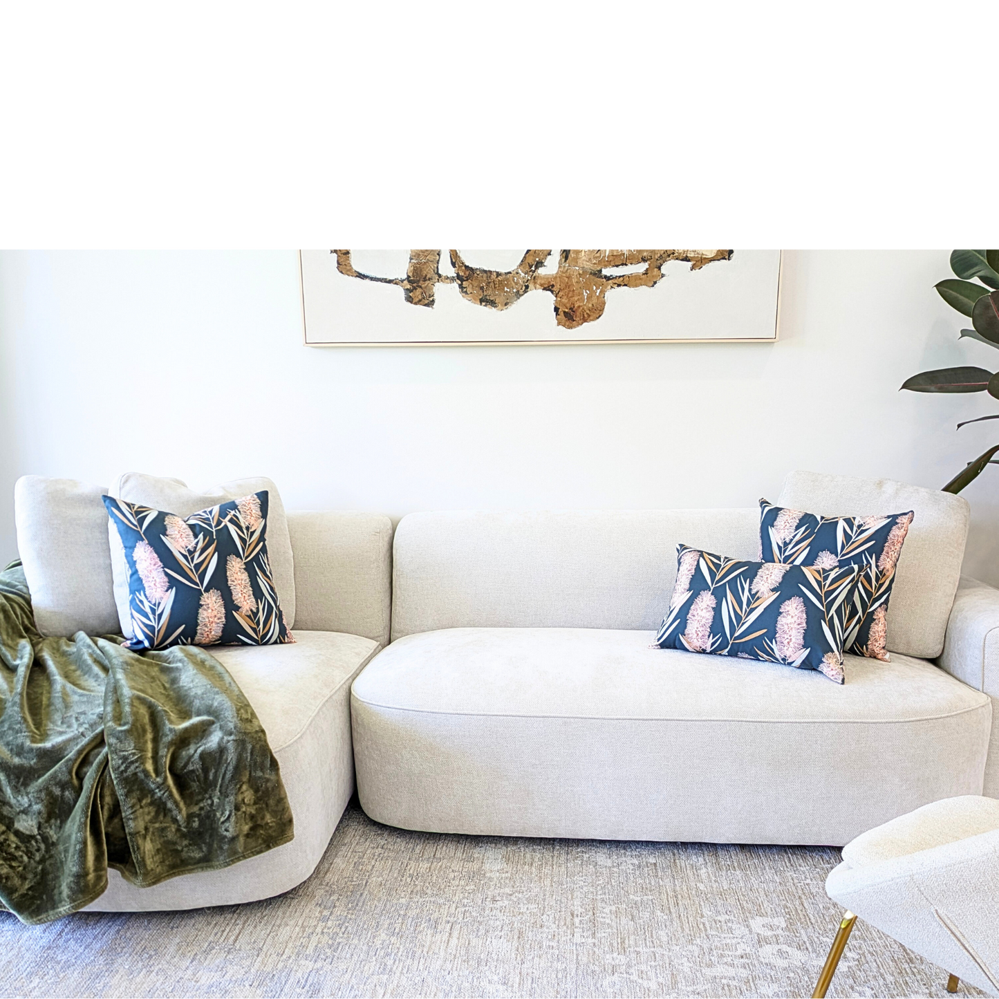 A modern living room with a light beige sectional sofa, two Emotive Collection Bottlebrush Cushion Covers in navy blue with pink pineapples (Jocelyn Proust design), a green throw blanket, large abstract wall art, and a leafy plant in the corner.