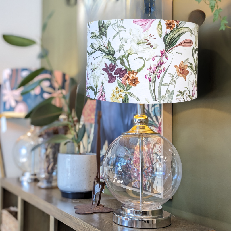 The Emotive Collection Summer Floral Lampshade tops a glass table lamp on a wooden shelf, surrounded by potted plants and decor, adding a cheerful touch to a stylish, well-lit room.