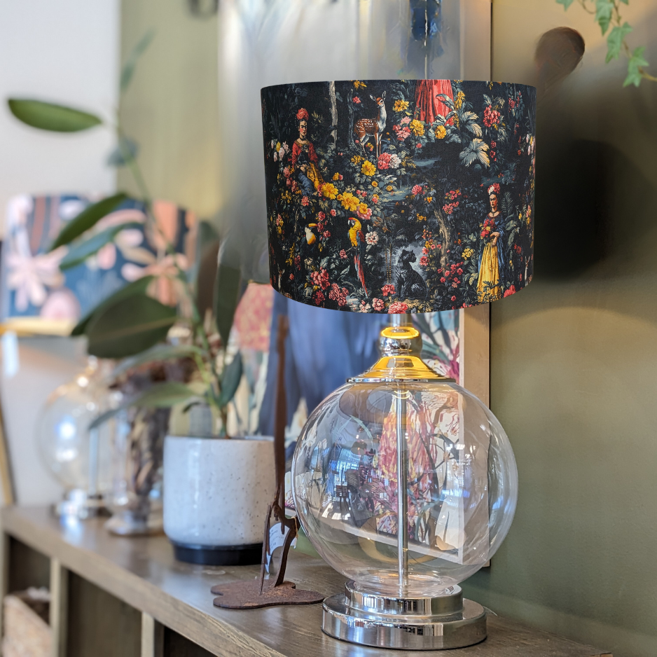 The Emotive Collection’s glass table lamp with a round base and Frida Kahlo Lampshade featuring a vibrant floral pattern sits on a wooden shelf beside potted plants and decor, with blurred artwork in the background.