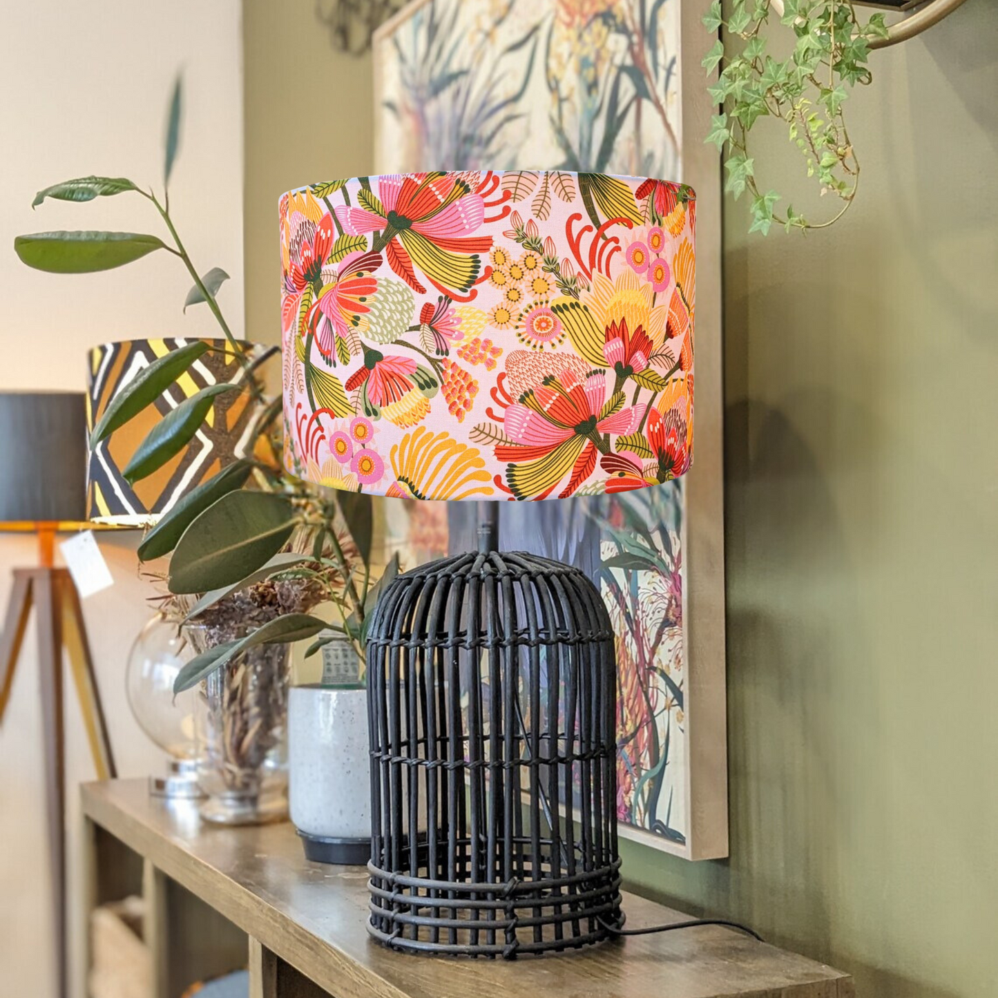 The Emotive Collection’s Wild Protea Lampshade with a black woven base sits on a wooden console table, surrounded by plants, decor, and framed art against a green wall. The handmade shade features modern botanical designs by Kirsten Katz.