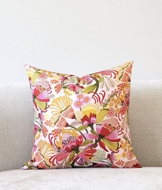 The Emotive Collection’s Wild Protea Cushion Cover displays Kirsten Katzs vibrant floral design in red, pink, yellow, and green on cotton canvas. It adds a pop of color to a light gray sofa set against a plain white wall.