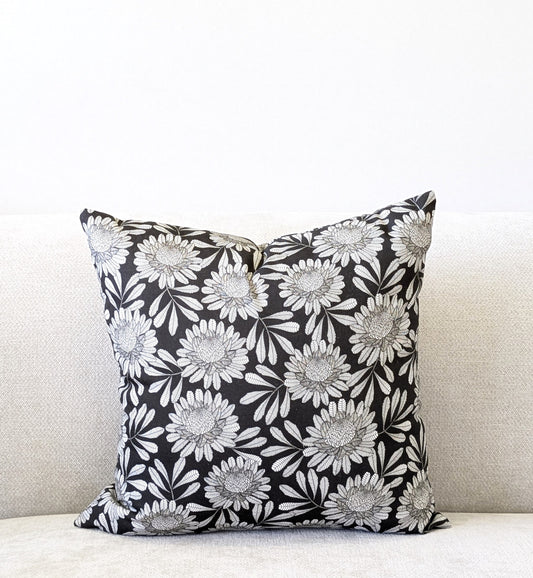 The Waratah Cushion Cover by Emotive Collection, showcasing a black base with white floral Jocelyn Proust design, rests on a light sofa against a plain white wall.