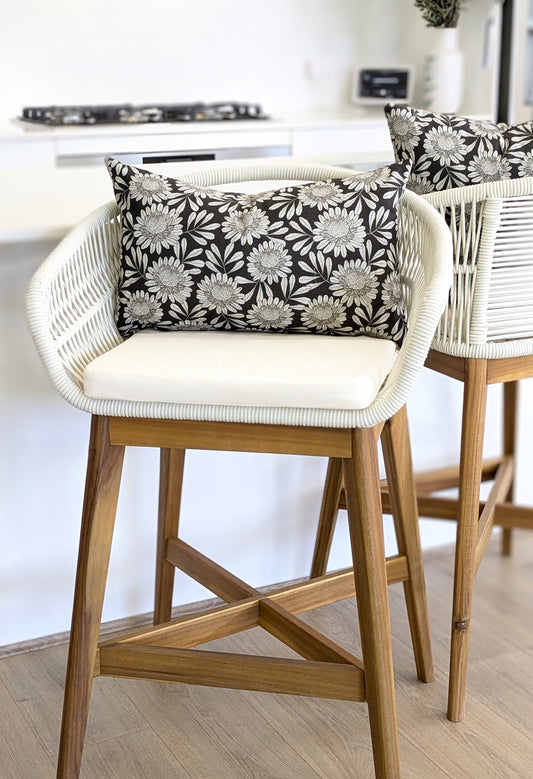 A modern wooden bar stool with a woven white frame, cream cotton canvas cushion, and a Waratah Cushion Cover by Emotive Collection in black-and-white floral design is by the countertop; another similar stool is partially visible.