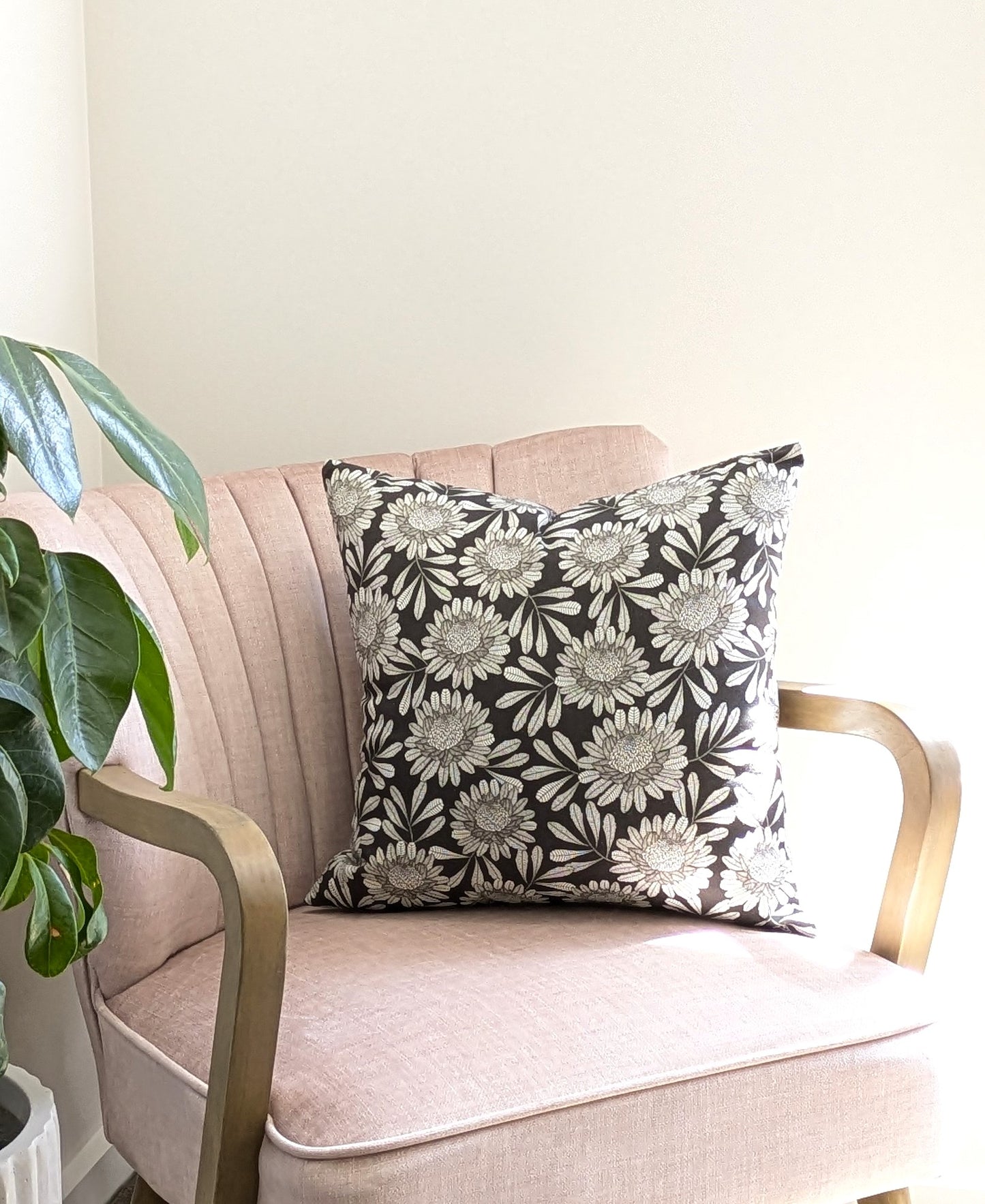 A blush pink armchair with gold armrests features the Waratah Cushion Cover by Emotive Collection, displaying a black and white floral pillow. A leafy green plant in a white pot sits nearby, all bathed in natural light.