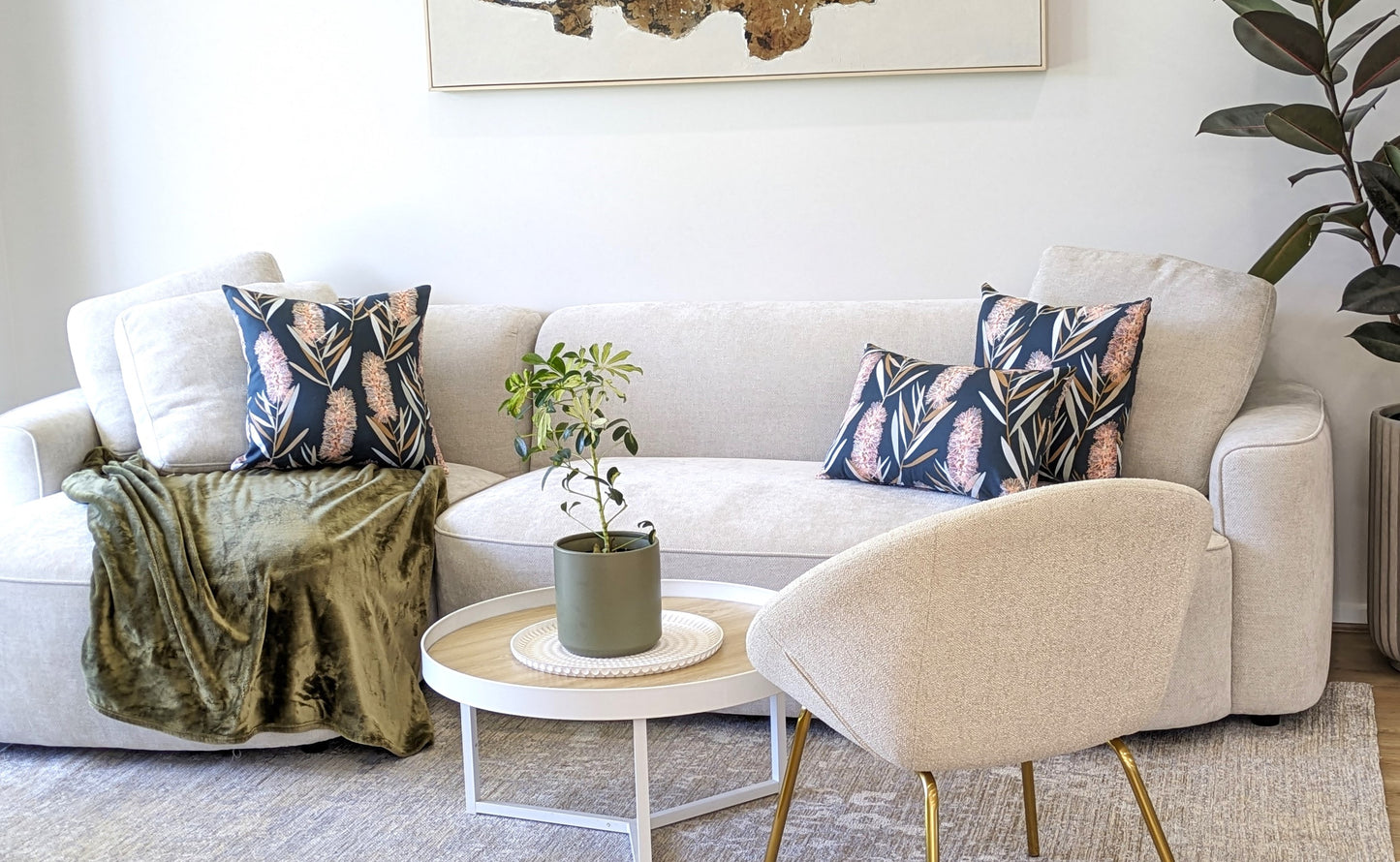 A modern living room featuring a light beige sofa, patterned throw pillows with a Blue Native Cushion Cover by Emotive Collection, green blanket, round coffee table with plant, cream armchair, and abstract wall art.
