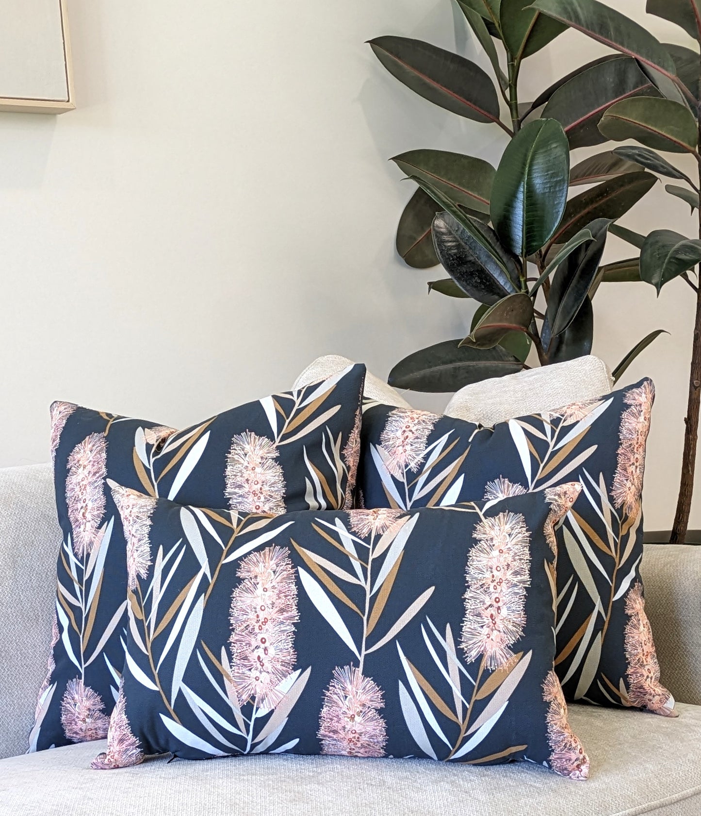 Three Emotive Collection Bottlebrush Cushion Covers with pink and white floral patterns are arranged on a light sofa. A leafy green plant behind the cushions enhances the cozy, modern setting.