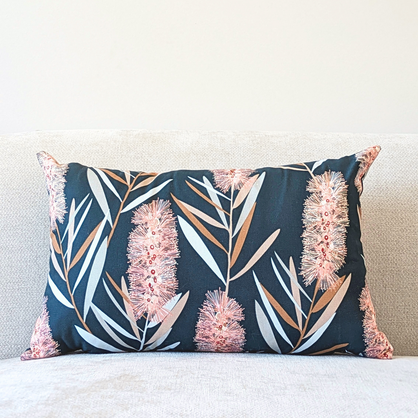 The Bottlebrush Cushion Cover from Emotive Collection is a rectangular handmade cotton canvas cover with Jocelyn Prousts pink bottlebrush flowers and light brown and gray leaves on a dark background, shown on a light-colored sofa.
