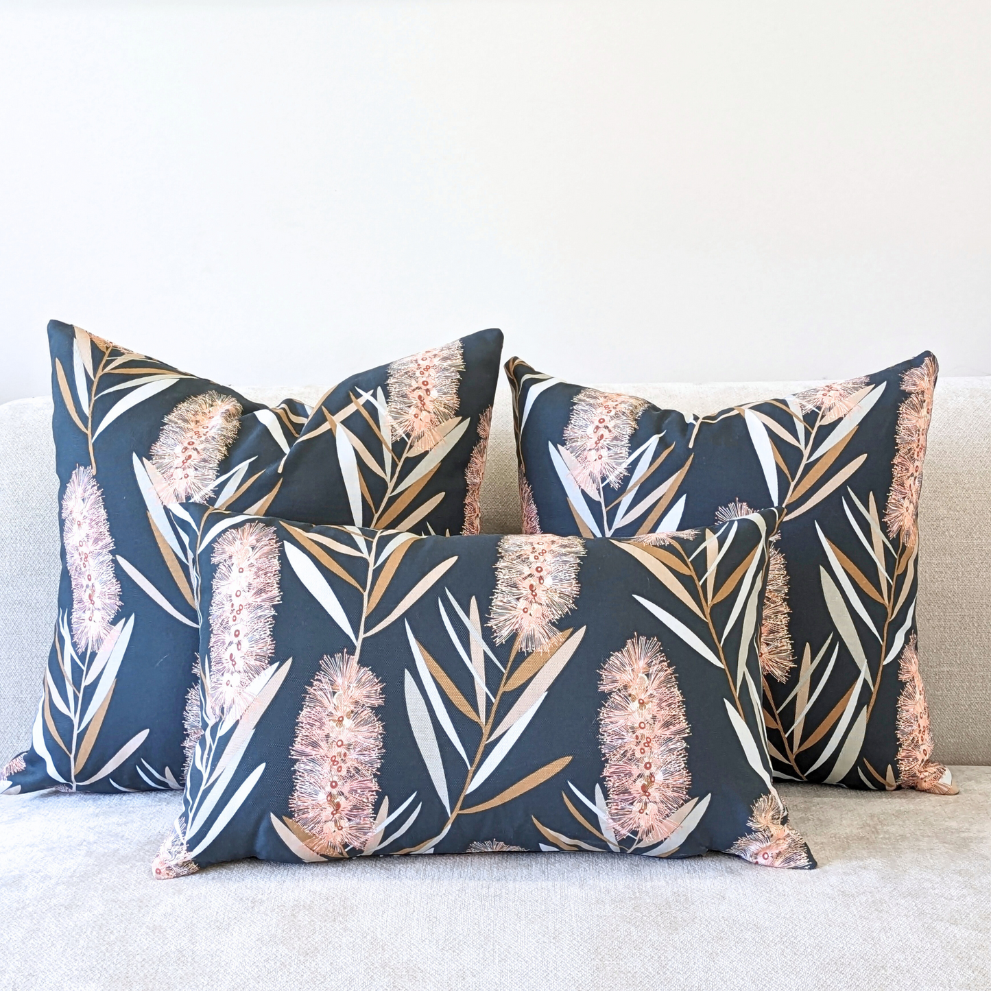Three Bottlebrush Cushion Covers by Emotive Collection, featuring floral and leaf patterns in light pink and beige on dark grey cotton canvas, are displayed on a light-colored sofa.