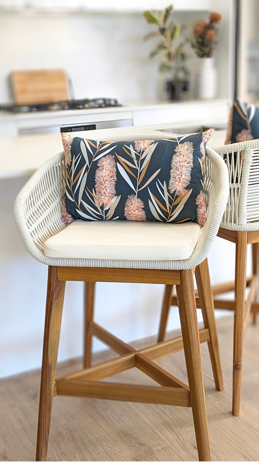 A modern barstool with a wooden frame and white woven seat is topped with an Emotive Collection Bottlebrush Cushion Cover in cotton canvas, featuring a Jocelyn Proust design. The stool sits at a white kitchen counter with blurred plants in the background.