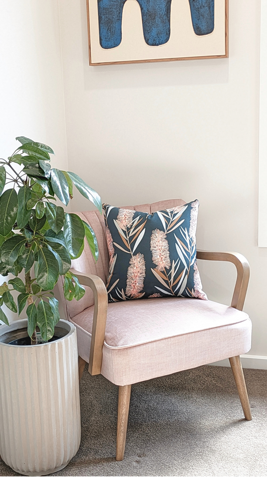 A light pink armchair with wooden arms holds the Bottlebrush Cushion Cover from Emotive Collection, featuring Jocelyn Proust designs. Nearby is a leafy plant in a ribbed white planter and abstract blue artwork hangs above the chair.
