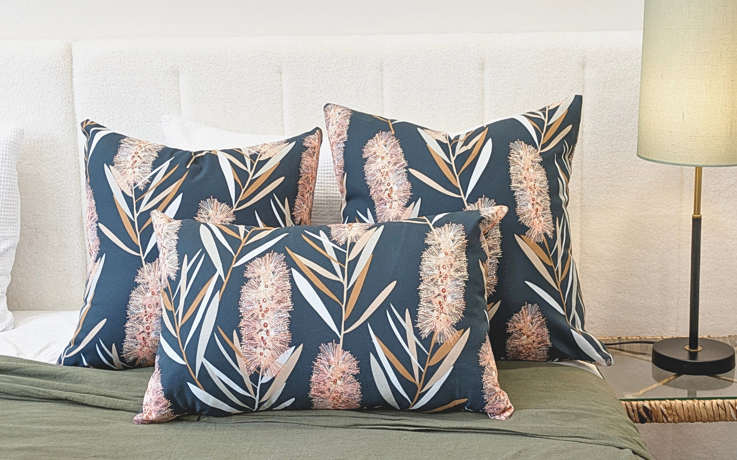 Three Bottlebrush Cushion Covers from the Emotive Collection, featuring a blue background and pink floral pattern on cotton canvas, rest on a bed with a white headboard, paired with a green blanket and a modern lamp on the bedside table.