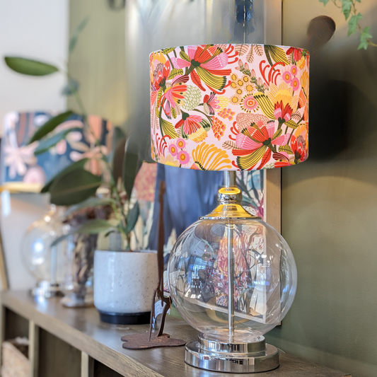 The Emotive Collection Wild Protea Lampshade, with its modern botanical design, sits on a wooden shelf among plants and decor, adding a vibrant yet cozy touch to the space.