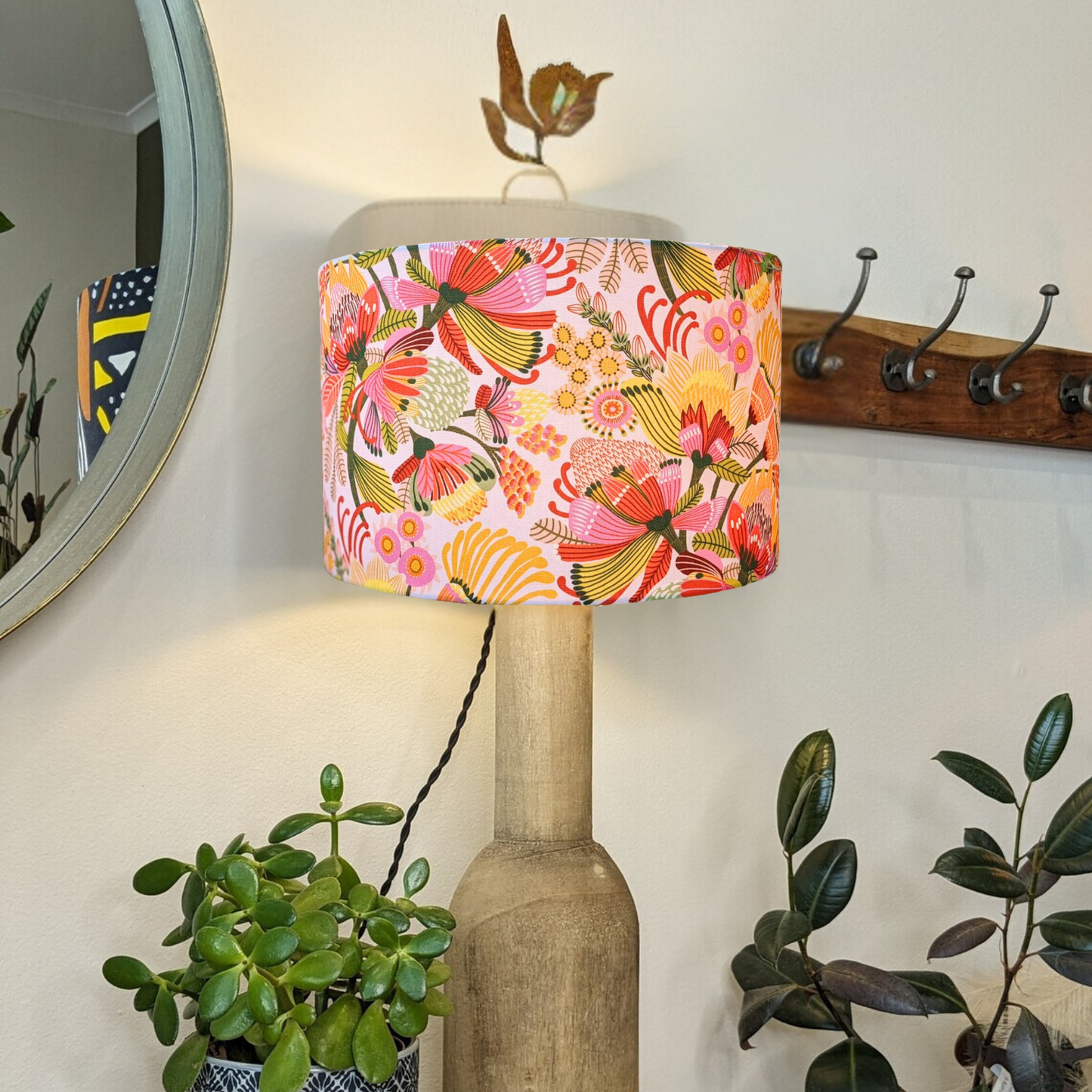 The Emotive Collection’s Wild Protea Lampshade, featuring modern botanicals, tops a wooden floor lamp beside a leafy potted plant. A round mirror and a wooden coat rack with hooks hang on the wall in the background.
