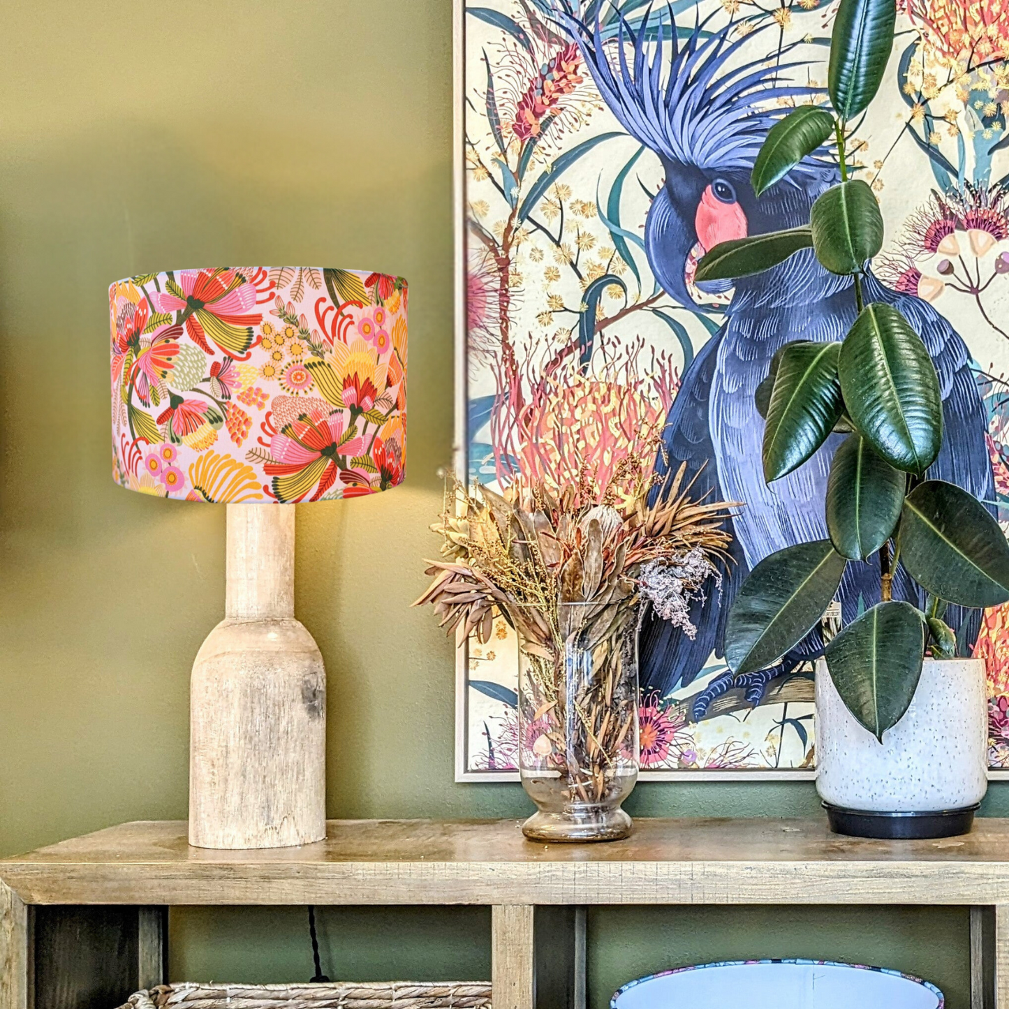 The Emotive Collection’s Wild Protea Lampshade, a wooden lamp with a modern botanical design by Kirsten Katz, sits on a shelf beside dried flowers, a potted rubber plant, and floral artwork of a black bird with red patch and spiky crest.
