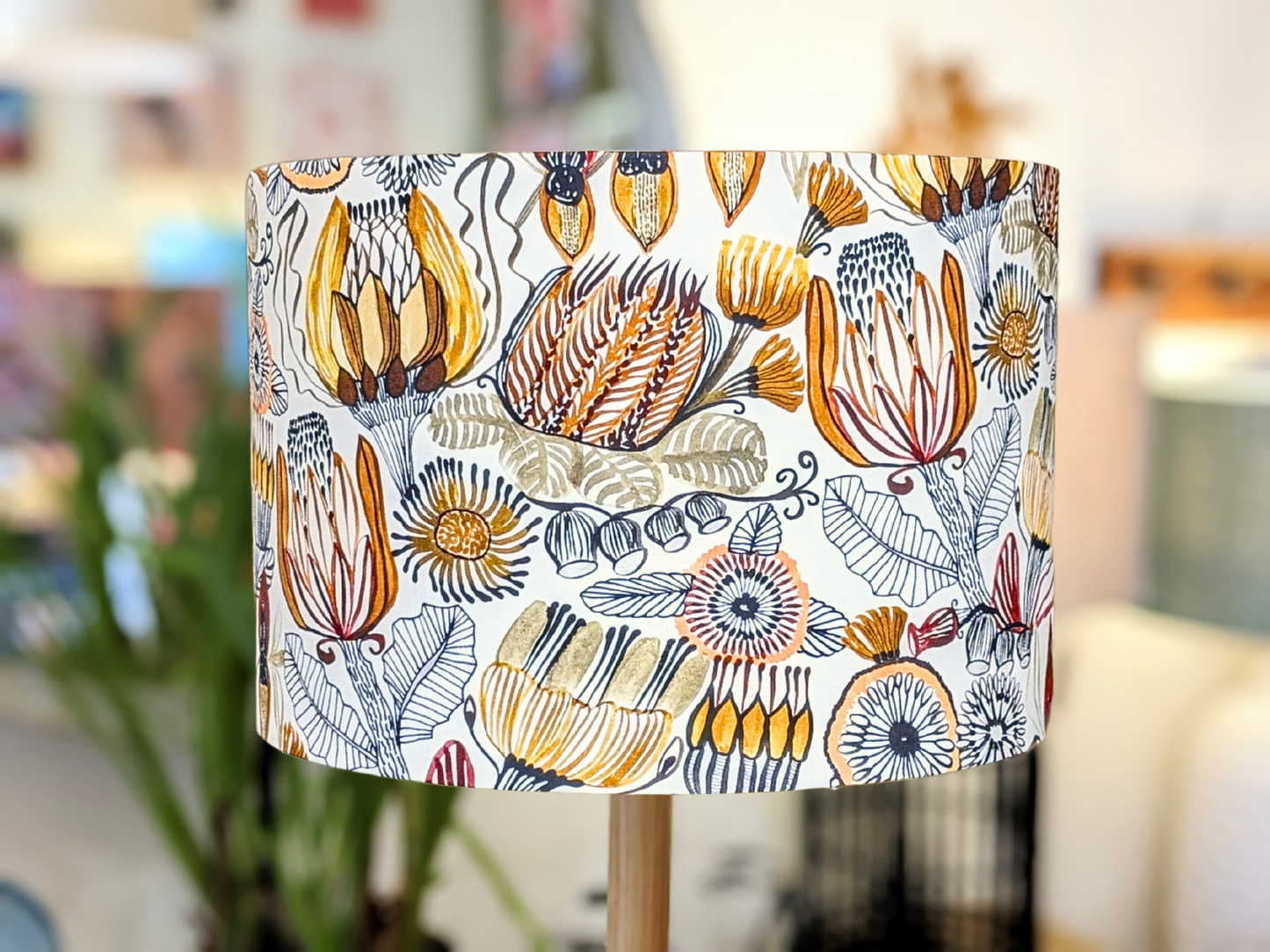 A close-up of the Emotive Collection’s Autumn Natives Lampshade, featuring colorful hand-drawn floral and leaf patterns in yellow, orange, brown, and gray, set against a blurred indoor background.