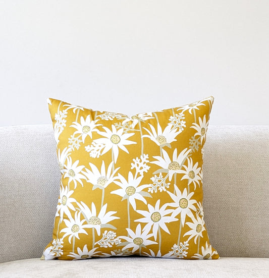 The Mustard Flannel Flower Cushion Cover from Emotive Collection, featuring a white daisy pattern inspired by Jocelyn Proust, adds a vibrant touch to a light beige couch against a plain white wall.