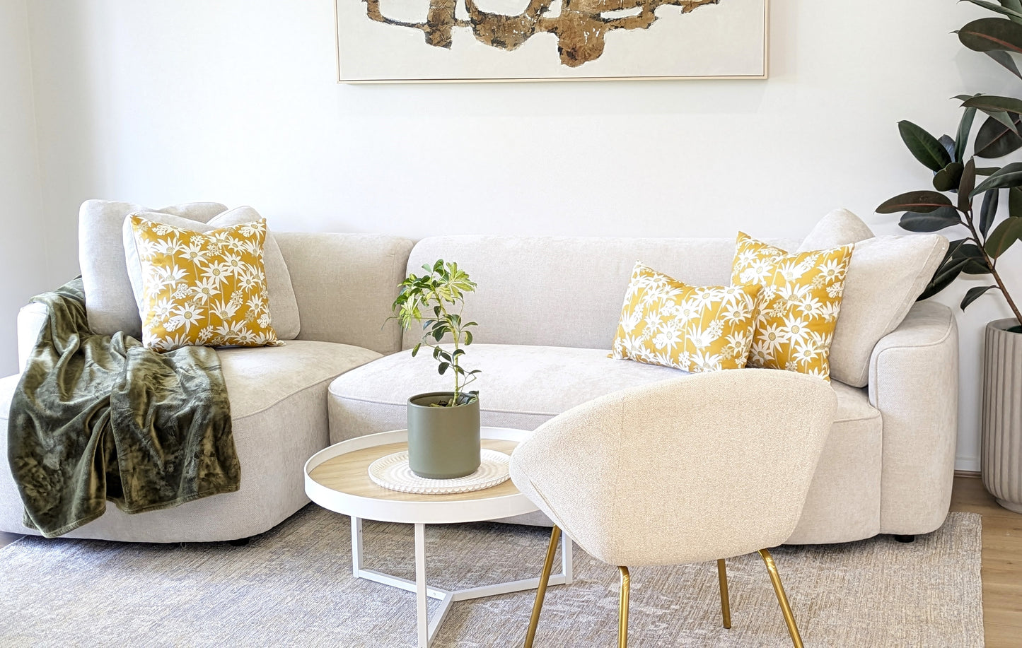 A modern living room with a light beige sofa, Emotive Collection’s Mustard Flannel Flower Cushion Covers, a green throw blanket, small round coffee table with potted plant, cream chair, large plant, and abstract wall art.