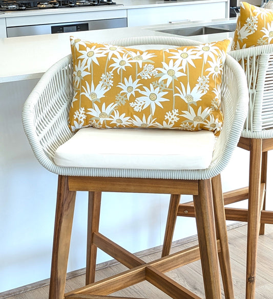A modern wooden barstool with a white woven backrest and seat cushion, styled with the Emotive Collections Mustard Flannel Flower Cushion Cover, designed by Jocelyn Proust, is shown in a bright kitchen setting.