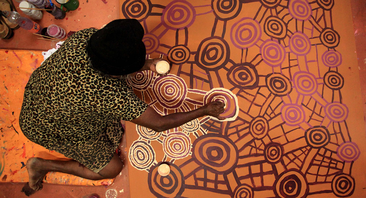 A person in a patterned dress sits on the floor, painting intricate circular patterns with earthy tones on a large canvas featuring traditional Aboriginal art designs. Paint containers are scattered nearby.