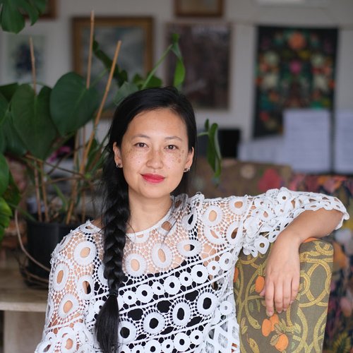 A woman with long black hair in a braid, wearing a white crochet top, sits indoors with her arm resting on a patterned chair. There are green plants and framed pictures in the background.