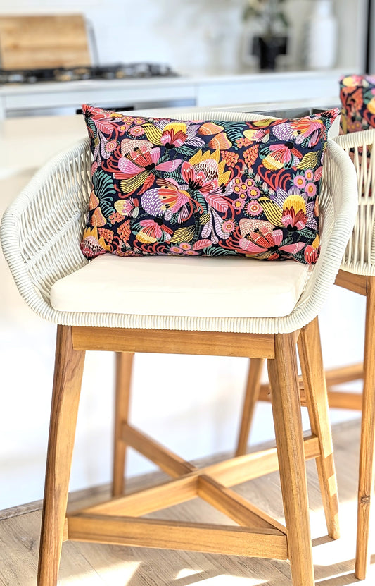 A wooden bar stool with a white woven backrest and seat cushion features an Emotive Collection Wild Protea Blue Cushion Cover, adding a floral accent in soft cotton canvas to the bright kitchen setting.