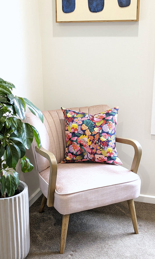 A pale pink armchair with wooden legs and arms is decorated with an Emotive Collection Wild Protea Blue Cushion Cover. A leafy plant in a white pot stands nearby, and abstract art by Kirsten Katz hangs above on the wall.