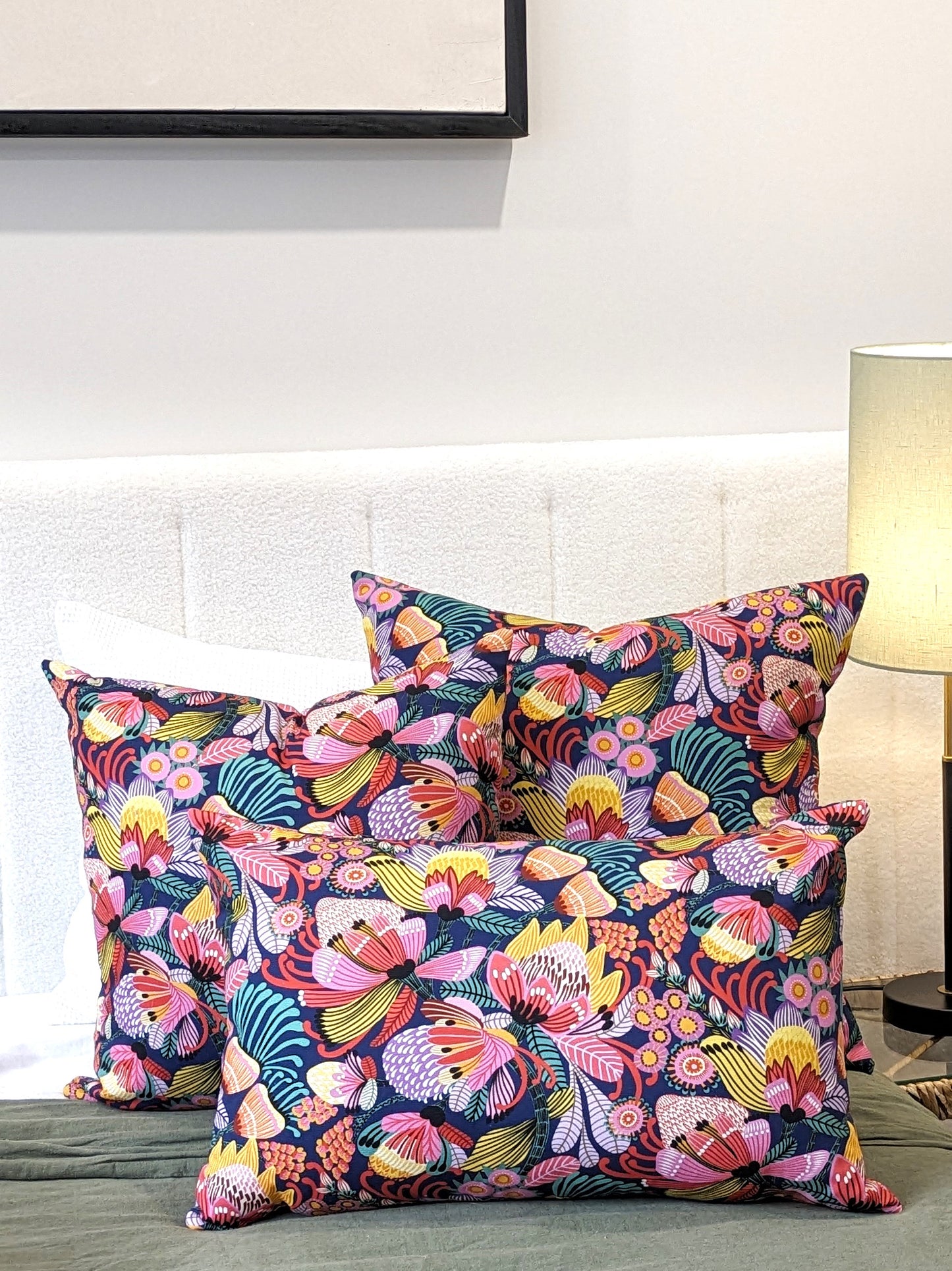 A bed features Emotive Collections Wild Protea Blue Cushion Cover by Kirsten Katz, arranged against a white upholstered headboard beside a lit lamp on the bedside table.