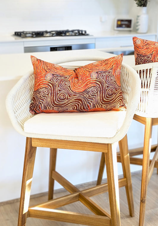 A white woven bar stool with a wooden frame features the Mina Mina Aboriginal Art Cushion Cover by Emotive Collection, showcasing an orange and brown abstract swirl. A similar set sits beside it in a modern, bright kitchen.