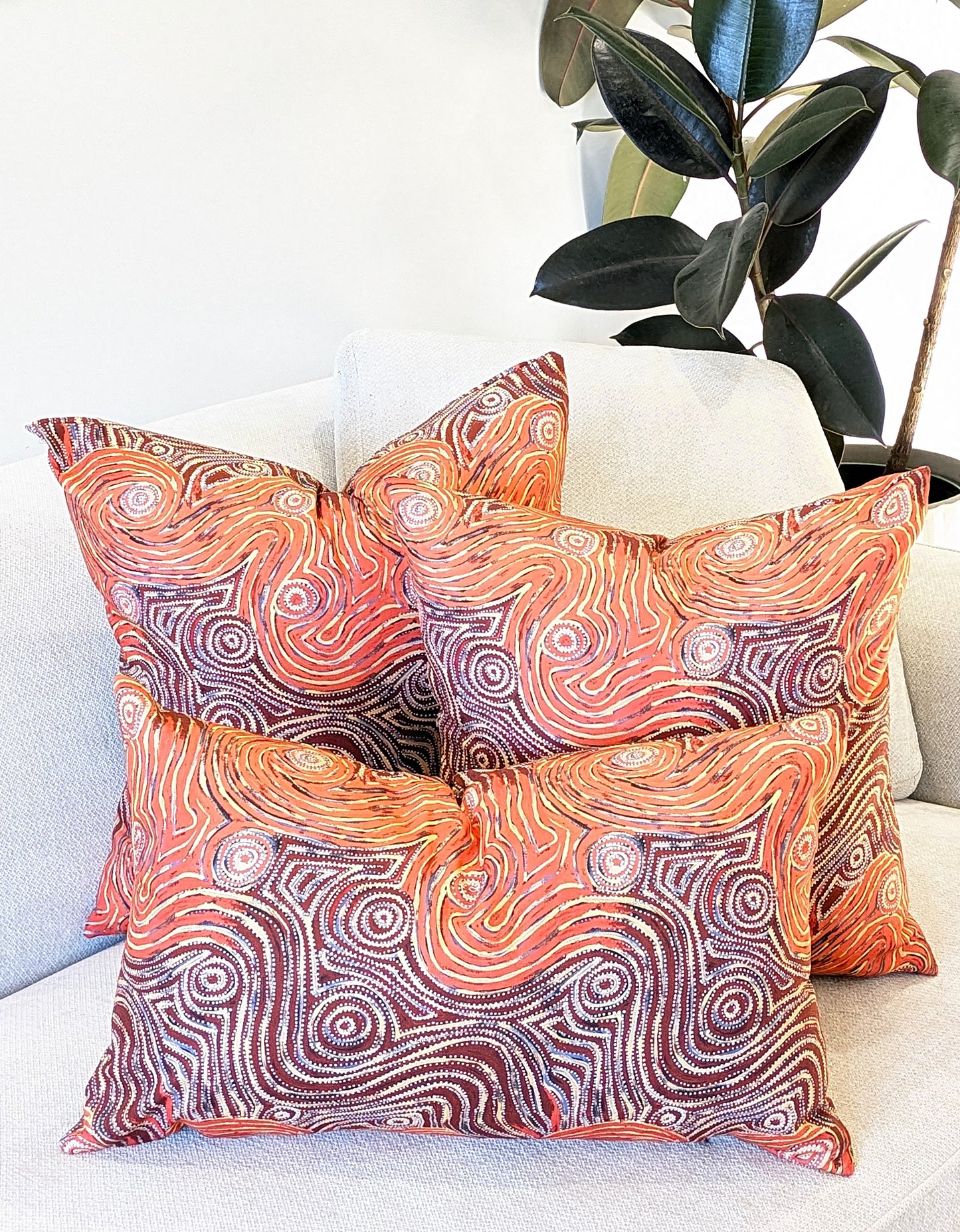 The Emotive Collection’s Mina Mina Aboriginal Art Cushion Cover showcases vibrant orange, white, and brown swirls on premium cotton canvas, displayed beside a leafy green plant.