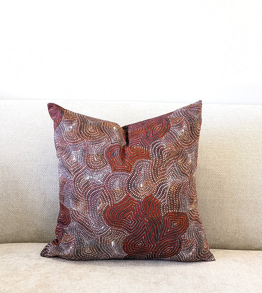 The Mina Mina Jukurrpa Aboriginal Art Cushion Cover by Emotive Collection, featuring abstract patterns in brown, red, and white, decorates a light-textured sofa set against a plain white background.
