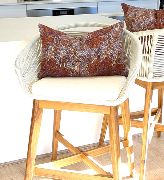 A modern barstool with a white woven back, wooden legs, and a handmade cushion is topped with an Emotive Collection Mina Mina Jukurrpa Aboriginal Art Cushion Cover in brown, white, and maroon. Another similar chair sits beside it.