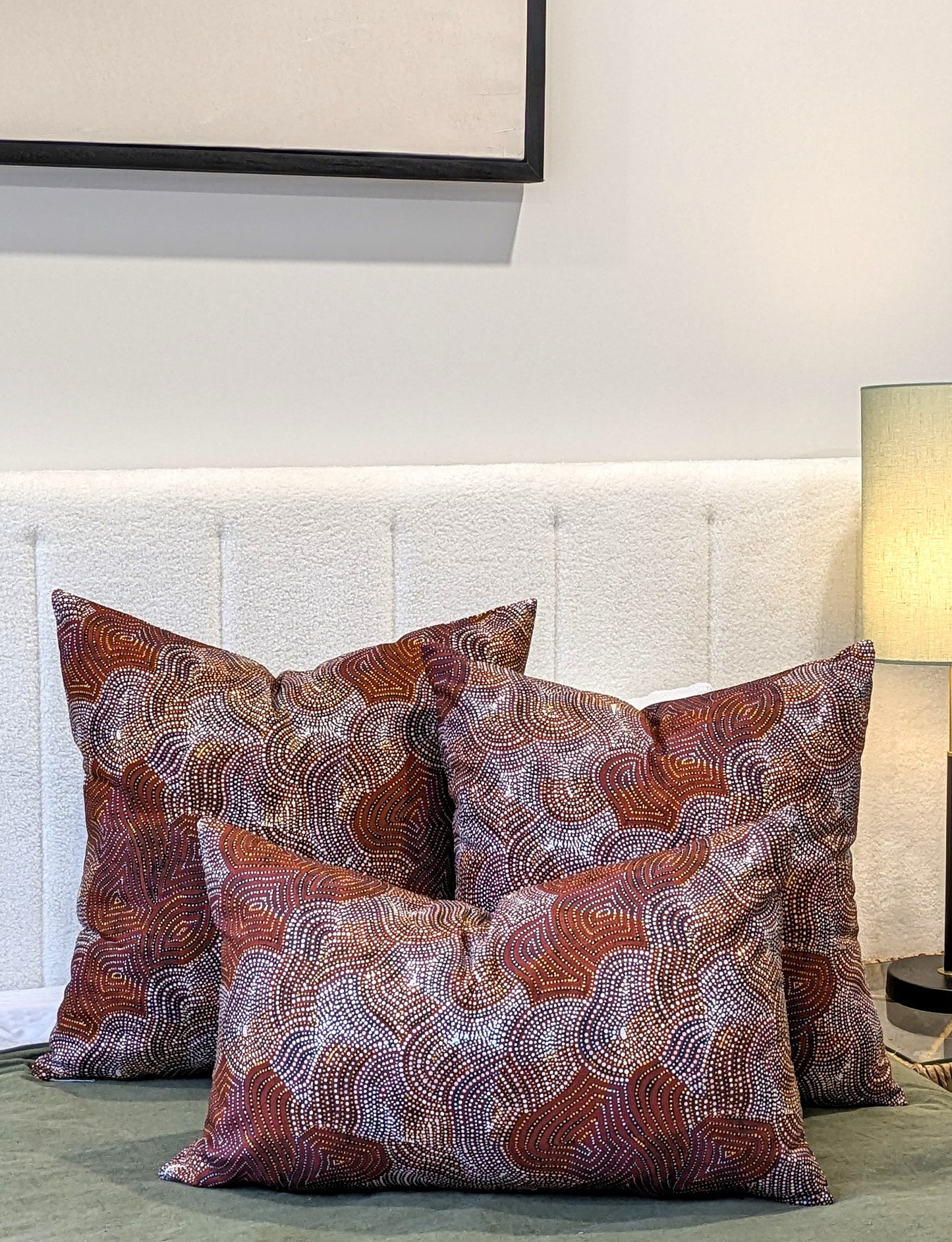 Three decorative pillows, including the Emotive Collection Mina Mina Jukurrpa Aboriginal Art Cushion Cover with wavy red, brown, and white patterns, rest on a bed with a white upholstered headboard. A lamp and framed picture add to the cozy setting.