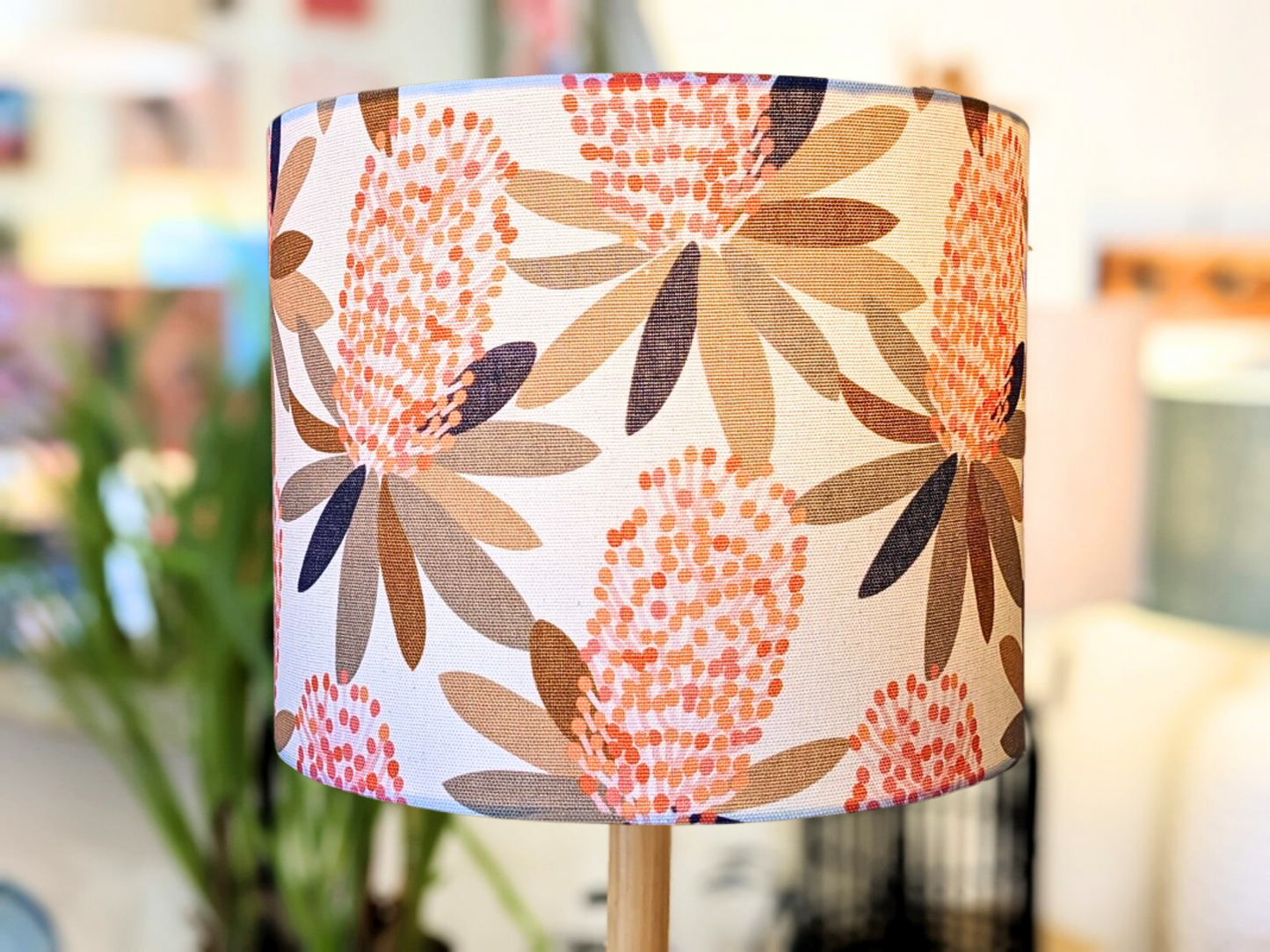 A close-up of the Emotive Collection’s Coastal Banksia Lampshade, featuring a botanical pattern with pink and orange flowers and brown leaves on a light background, inspired by Jocelyn Proust designs, set in a softly blurred indoor scene.