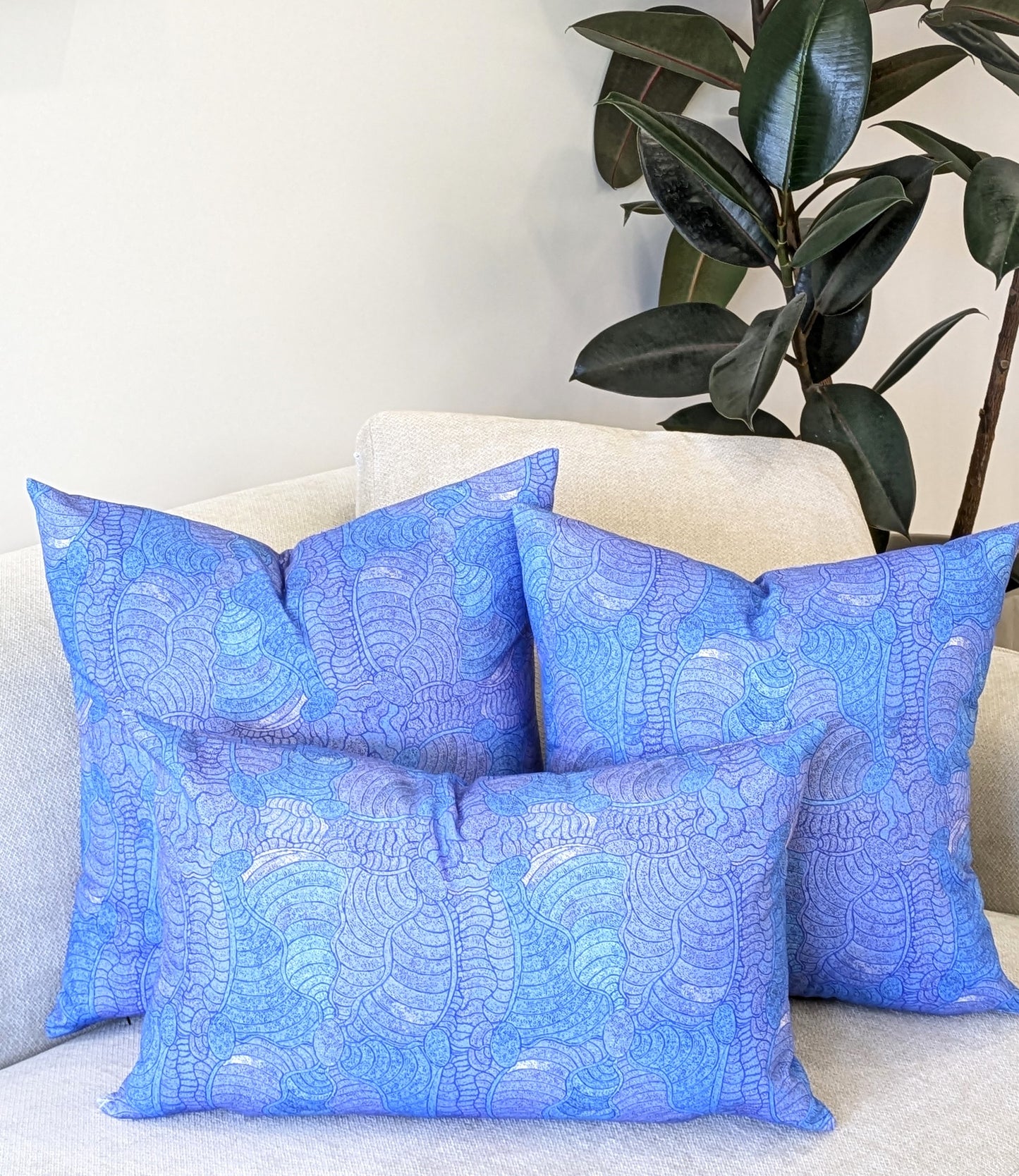 Four blue decorative pillows, including the Emotive Collection's Brushtail Possum Aboriginal Art Cushion Cover, decorate a beige sofa. A leafy rubber plant adds greenery to the neutral room.