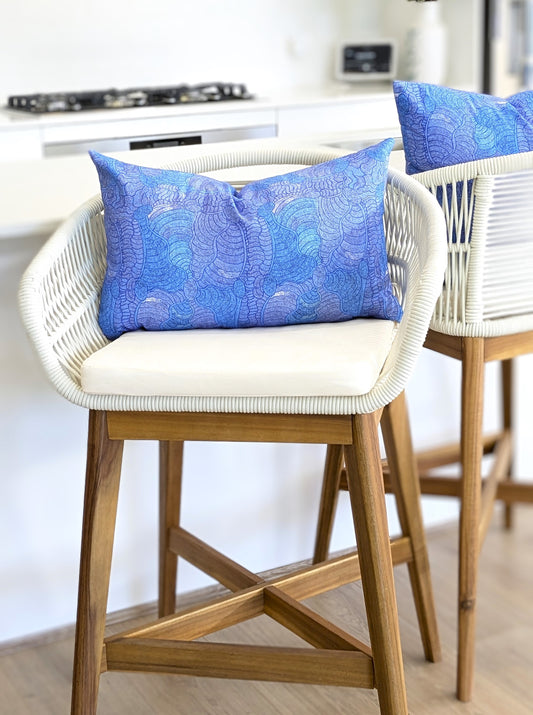 A white wicker and wood barstool with a premium cotton canvas cushion sits at the counter, topped with the Emotive Collection's blue Brushtail Possum Aboriginal Art Cushion Cover, inspired by an Indigenous Aboriginal artist.