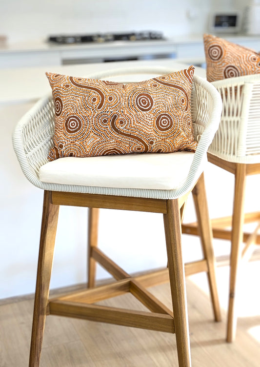 A white wicker bar stool with wooden legs and a white cushion features the Emotive Collection's Goanna Dreaming Aboriginal Art Cushion Cover, showcasing an abstract circular design by an Indigenous artist. Another stool is blurred in the background.