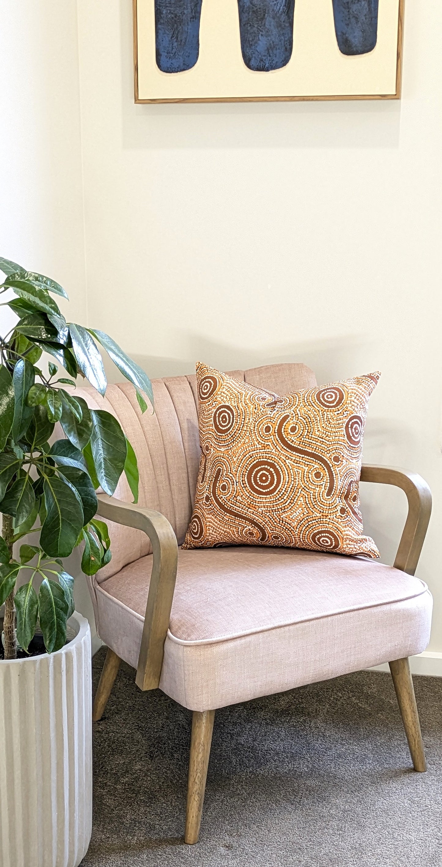 A light pink armchair with wooden arms holds the Emotive Collection’s Goanna Dreaming Aboriginal Art Cushion Cover. A leafy plant in a ribbed pot sits beside, with Indigenous abstract art displayed above.