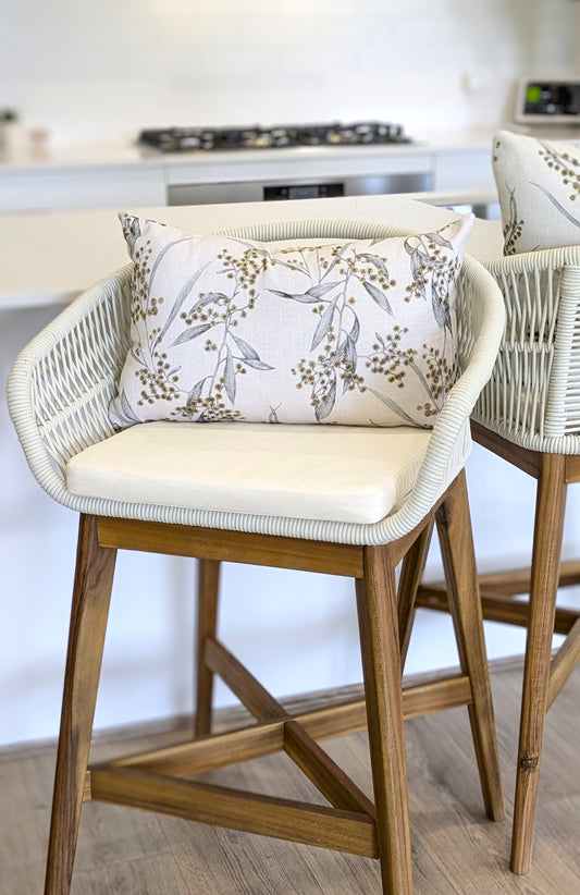 A modern wicker and wood barstool with a cream cushion and an Emotive Collection Golden Wattle Cushion Cover sits in a bright kitchen, with another similar stool nearby.