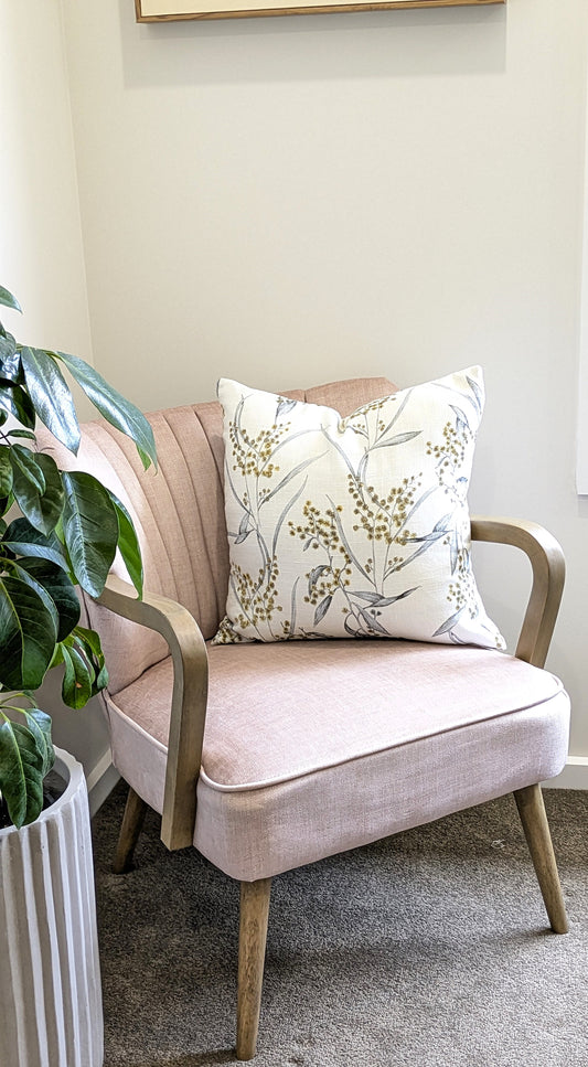 A pale pink cotton-linen chair with wooden legs holds a Golden Wattle Cushion Cover from Emotive Collection. The chair rests on a gray carpet next to a tall potted green plant in a ribbed pot.