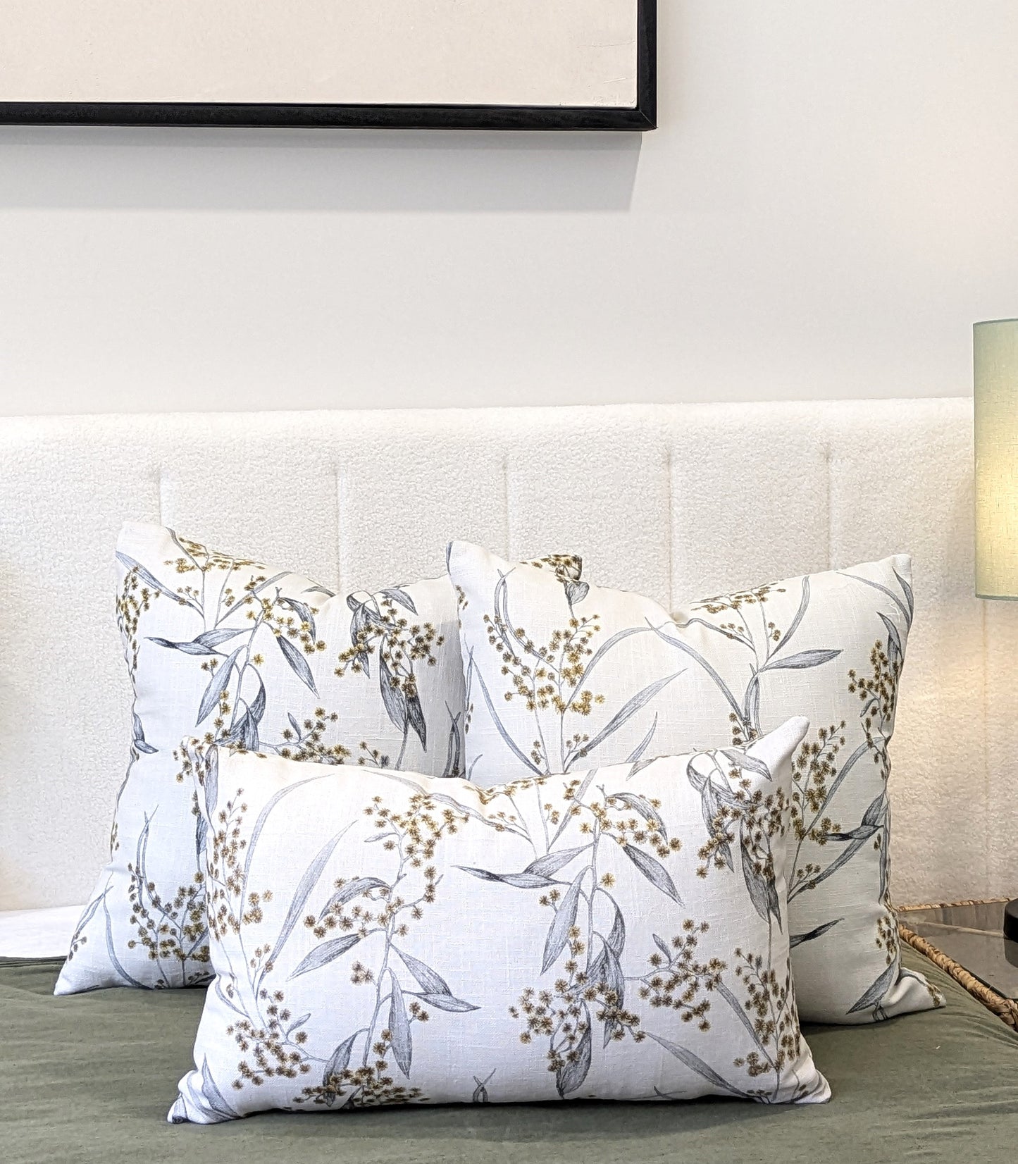 Three Emotive Collection Golden Wattle Cushion Covers, crafted from cotton linen with a botanical pattern, are arranged on a light headboard. Their green accents coordinate with the lamp and framed artwork in the background.