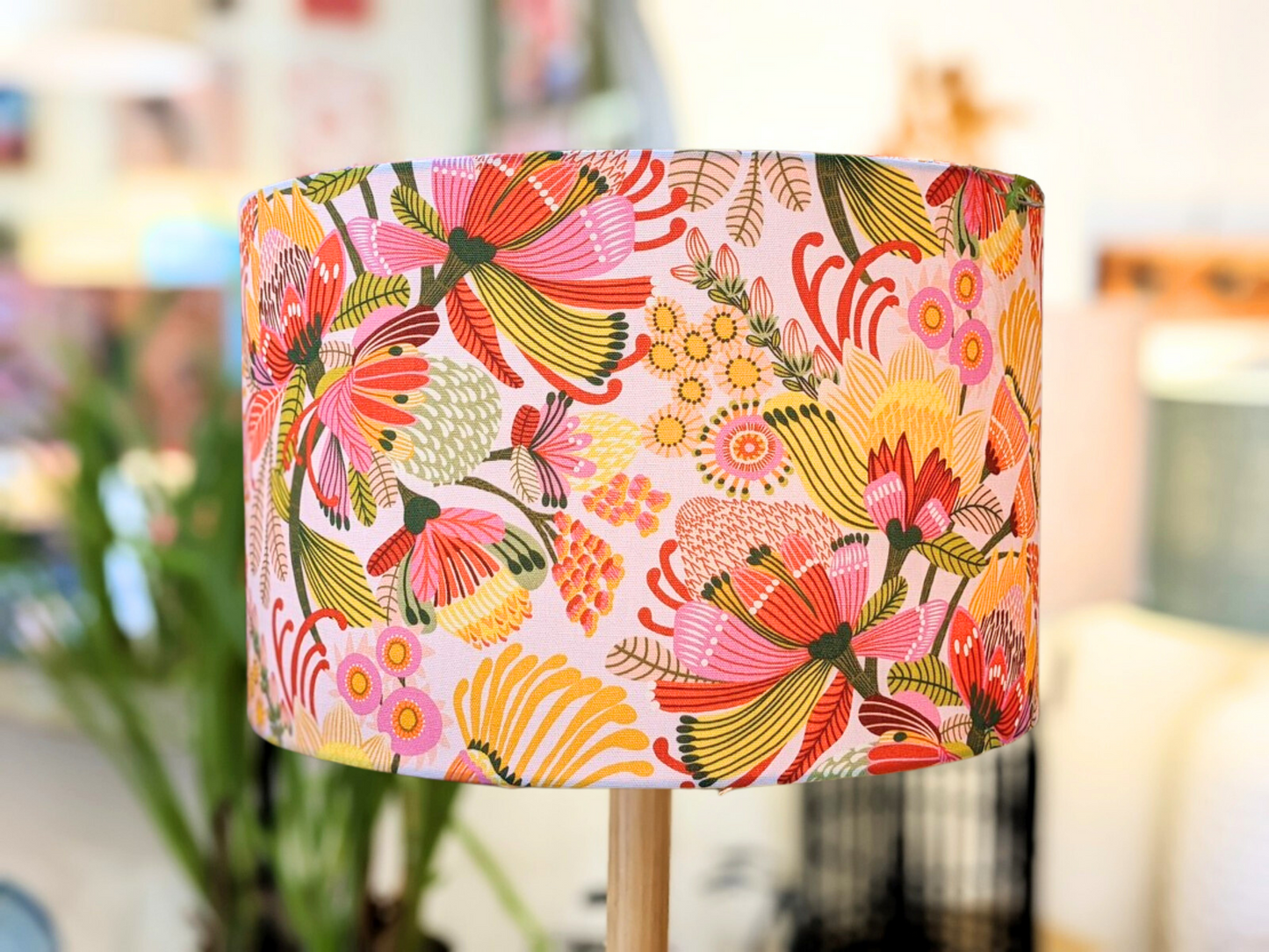 The Wild Protea Lampshade from Emotive Collection showcases Kirsten Katz’s modern botanicals—a handmade piece with a vibrant floral pattern in red, pink, yellow, and green on a softly blurred background.