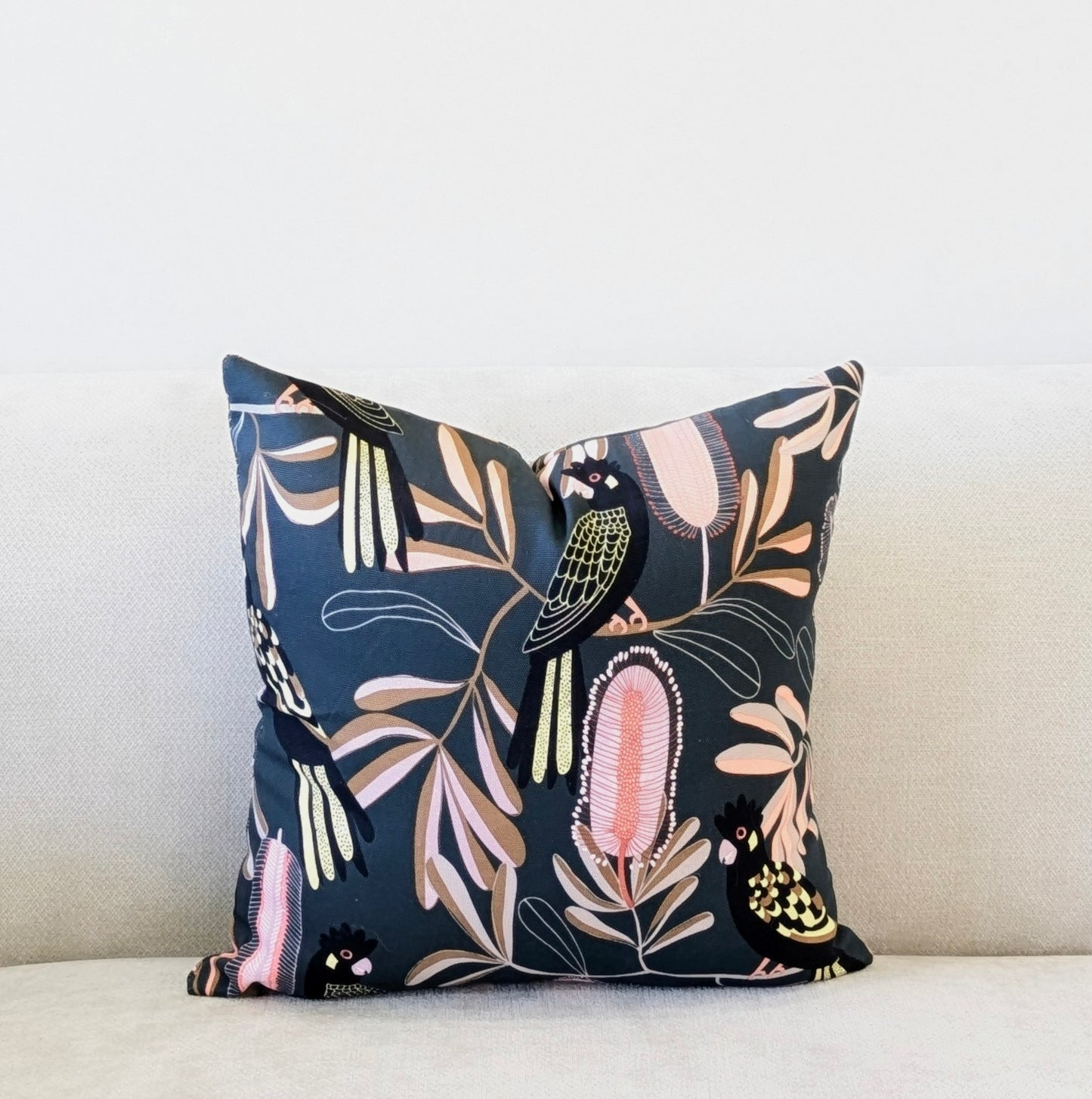 An Emotive Collection Yellow-Tailed Black Cockatoo Cushion Cover, showcasing Jocelyn Prousts vibrant design, adds a stylish touch to a light beige sofa set against a plain white wall.