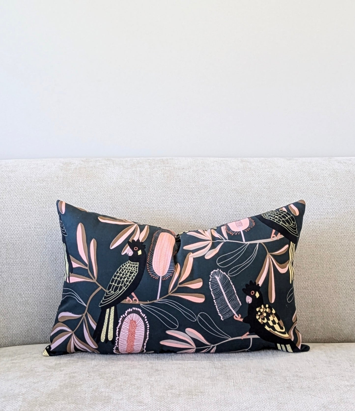 The Emotive Collection Yellow-Tailed Black Cockatoo Cushion Cover, with Jocelyn Prousts floral and bird design in pink, cream, and yellow cotton canvas, sits on a light beige sofa.