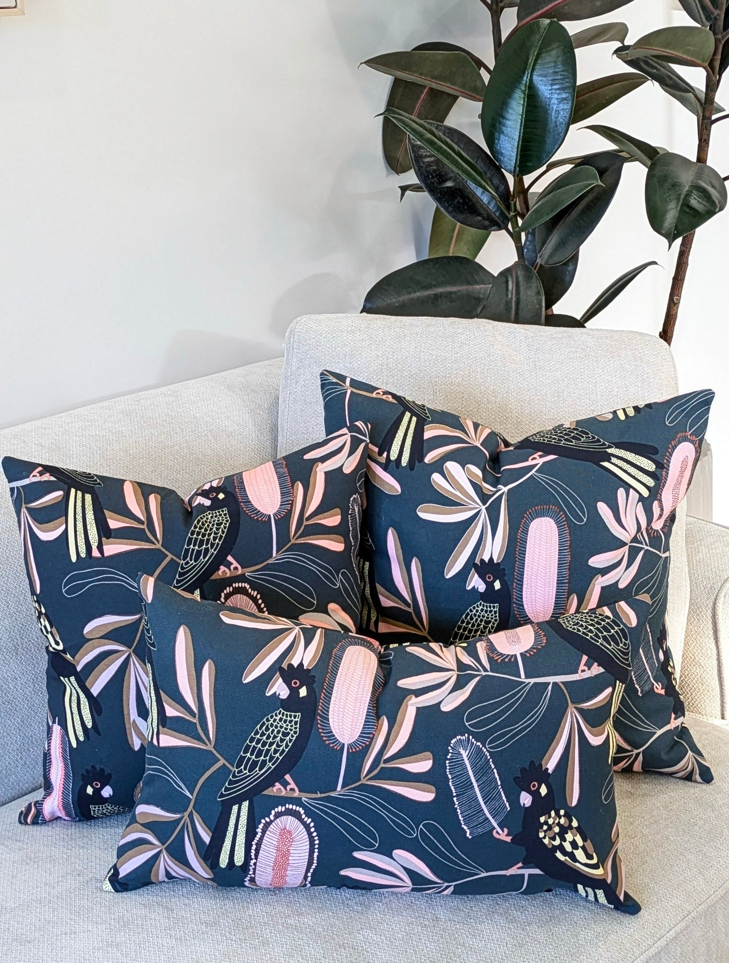 Three Emotive Collection Yellow-Tailed Black Cockatoo Cushion Covers, featuring pastel botanical designs by Jocelyn Proust, are arranged on a light armchair beside a tall green plant.