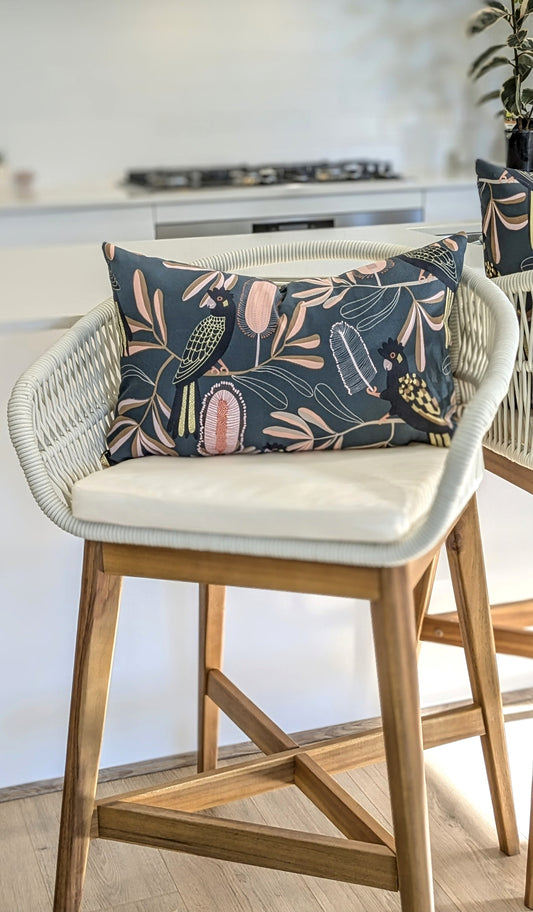 A modern white and wood bar stool is topped with the Emotive Collection Yellow-Tailed Black Cockatoo Cushion Cover, adding a vibrant accent to a bright kitchen setting.