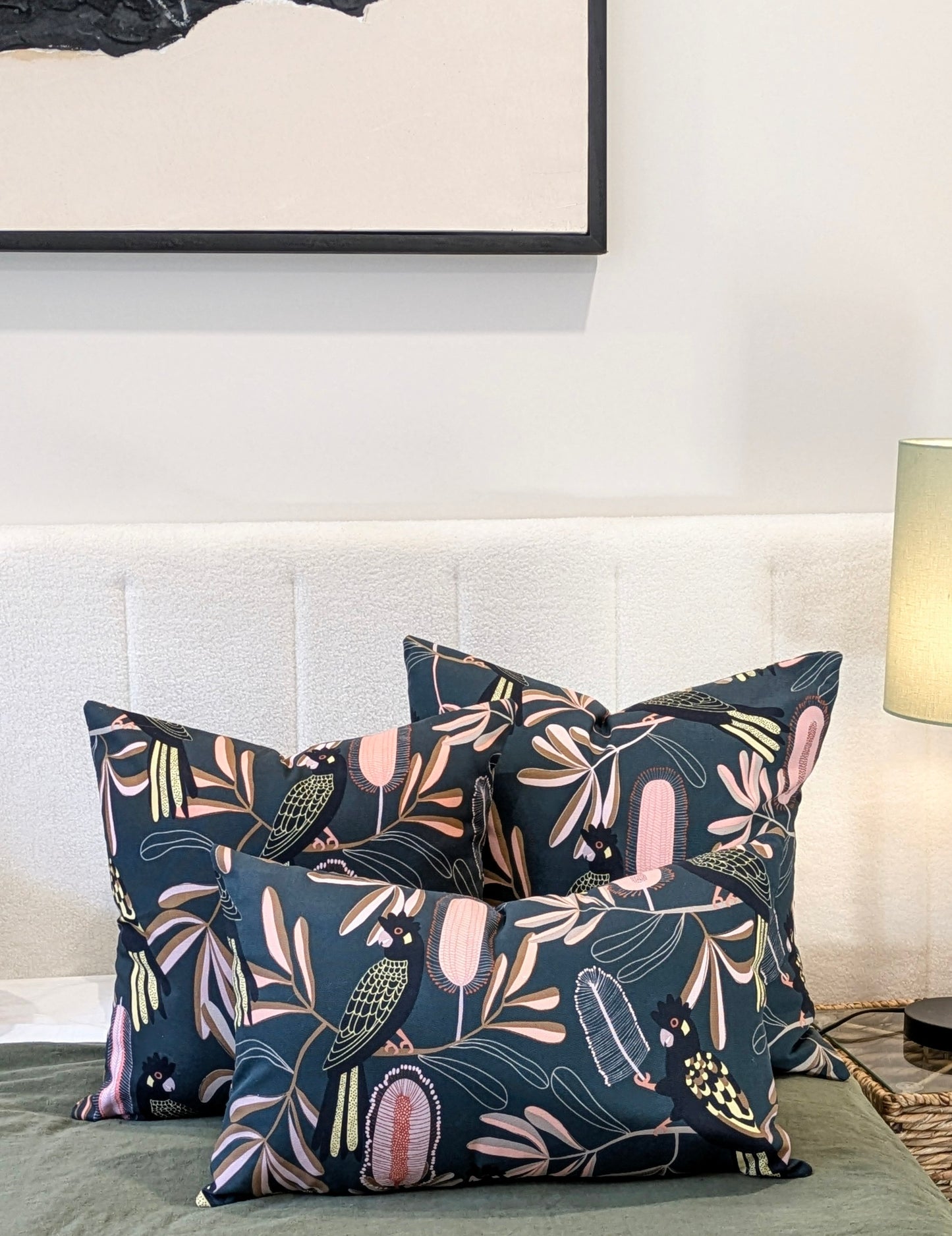 Three Emotive Collection Yellow-Tailed Black Cockatoo Cushion Covers rest on a white upholstered headboard. A green blanket, a lit table lamp, and framed abstract artwork complete the scene.
