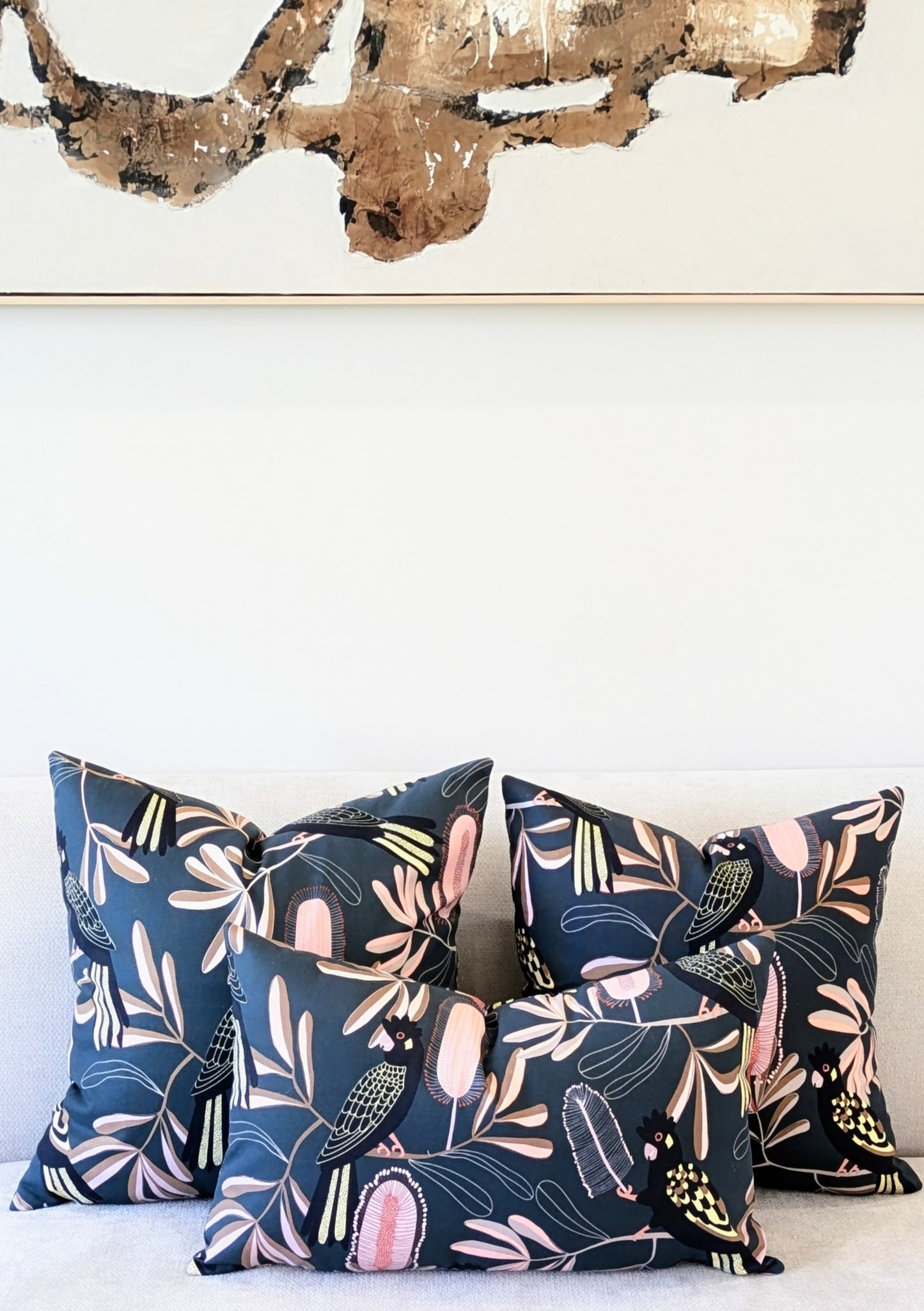 Four Emotive Collection Yellow-Tailed Black Cockatoo Cushion Covers with abstract leaf and bird patterns decorate a light sofa. Above, a gold and brown modern art painting completes the look.