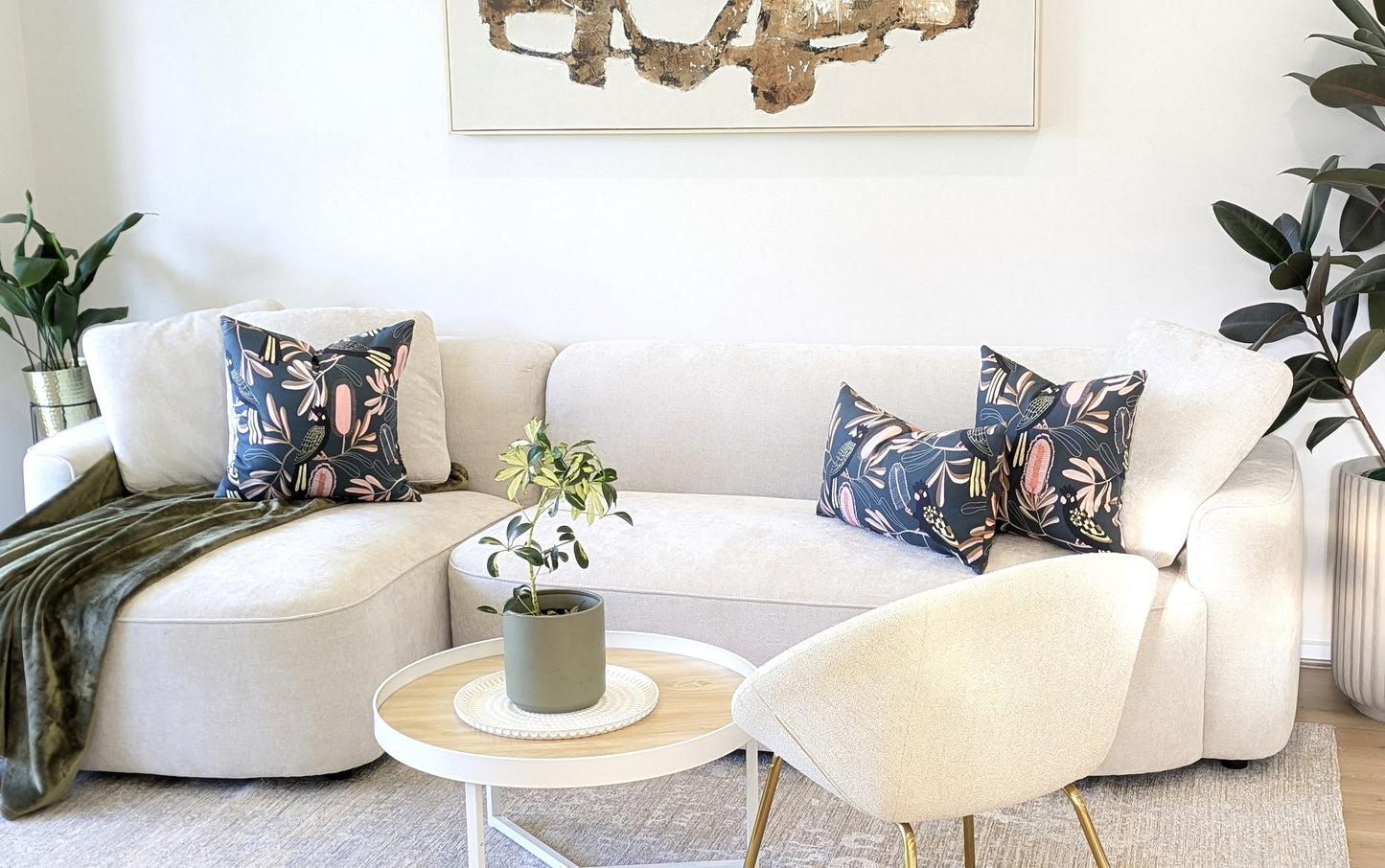 A modern living room with a white sectional sofa, three Emotive Collection Yellow-Tailed Black Cockatoo cushion covers, a green throw, round white coffee table with potted plant, cream armchair, and abstract wall art above the sofa.