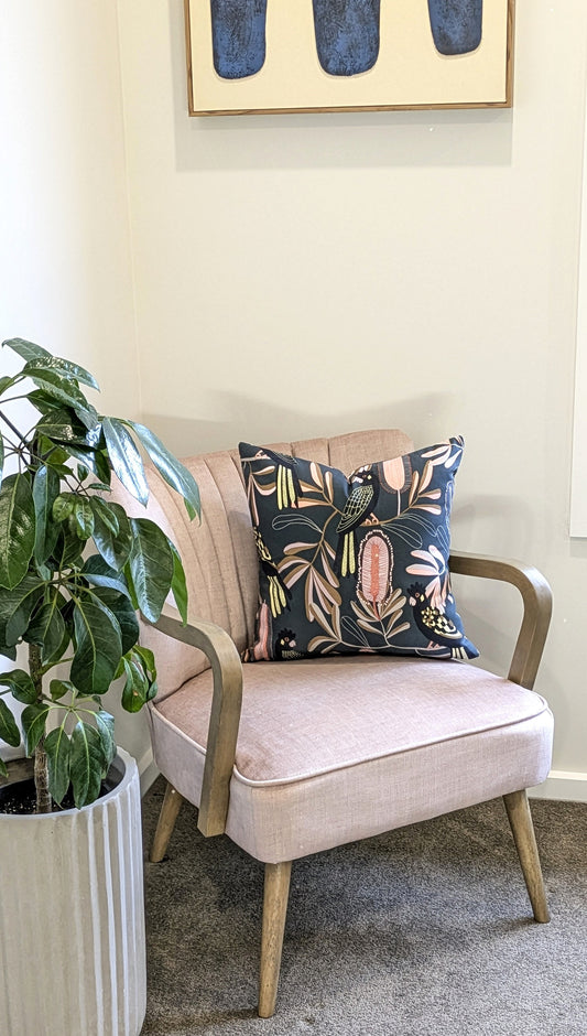 A light pink armchair with wooden arms sits in a corner, topped with the Emotive Collections Yellow-Tailed Black Cockatoo Cushion Cover. A green leafy potted plant is to the left, and abstract wall art hangs above.