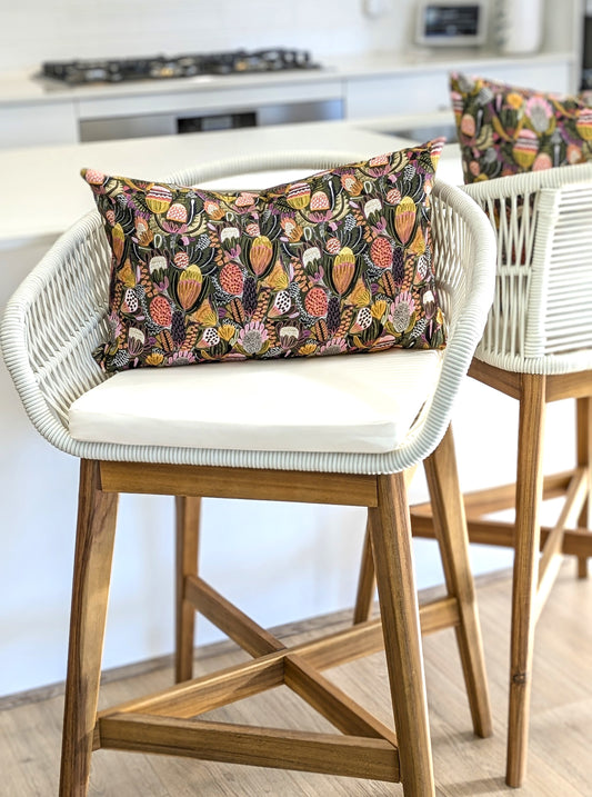 A white bar stool with a wooden base and woven backrest, topped with an Emotive Collection Bush Banksia Cushion Cover, stands in a bright kitchen next to a matching stool.